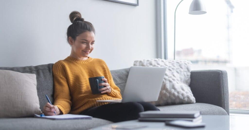 A woman abiding by financial rules sits on a couch with a laptop and a cup of coffee.