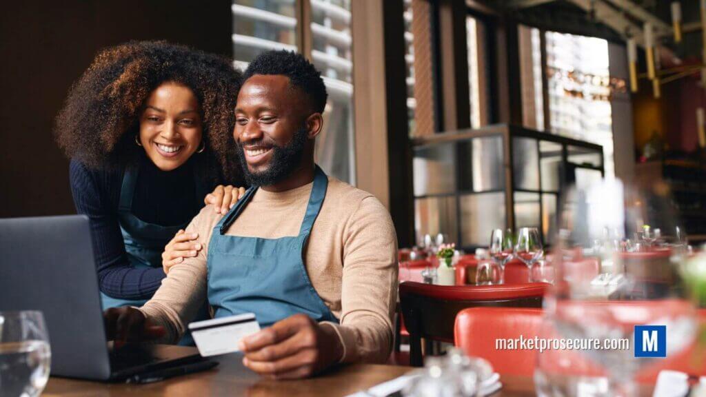 Small Business Owners Shopping Online with Credit Card A man, wearing an apron, sits at a restaurant table with his laptop and credit card beside an RFID wallet. A woman stands next to him, smiling at the screen. The restaurant setting is serene with empty tables and chairs. Visible in the scene is "marketprosecure.com." Ideal for your personal finance website’s visual content strategy!.